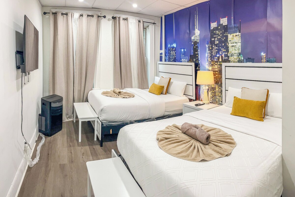 Une chambre d’hôtel avec deux lits, un bureau, une chaise, un téléviseur et une fresque représentant un paysage urbain sur le mur.