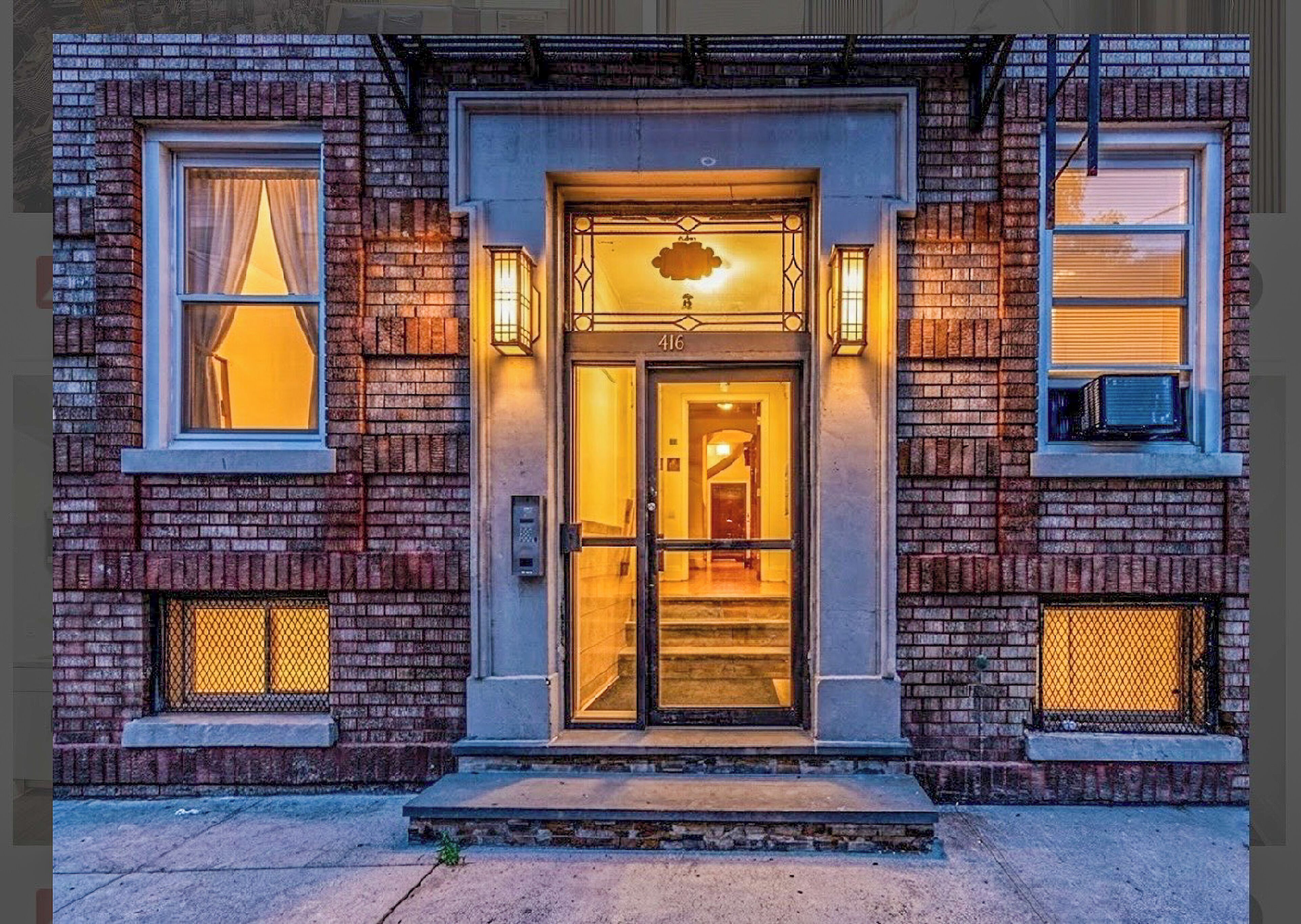 A brick building with a numbered door, two windows with curtains, and a small porch area.