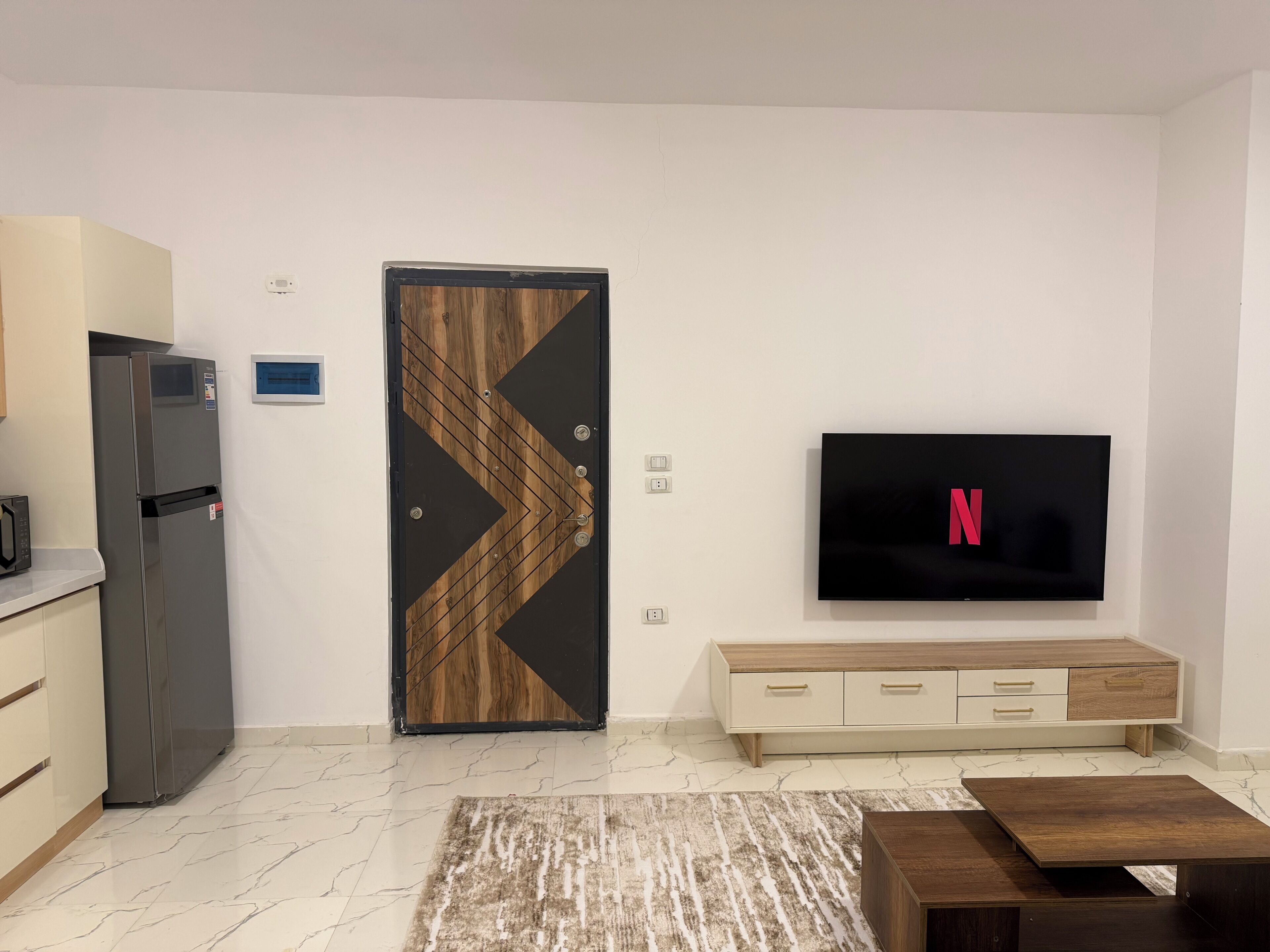 Una sala de estar moderna con una televisión de pantalla plana, una puerta de madera con diseño geométrico, un mueble empotrado para la TV y un piso de mármol.