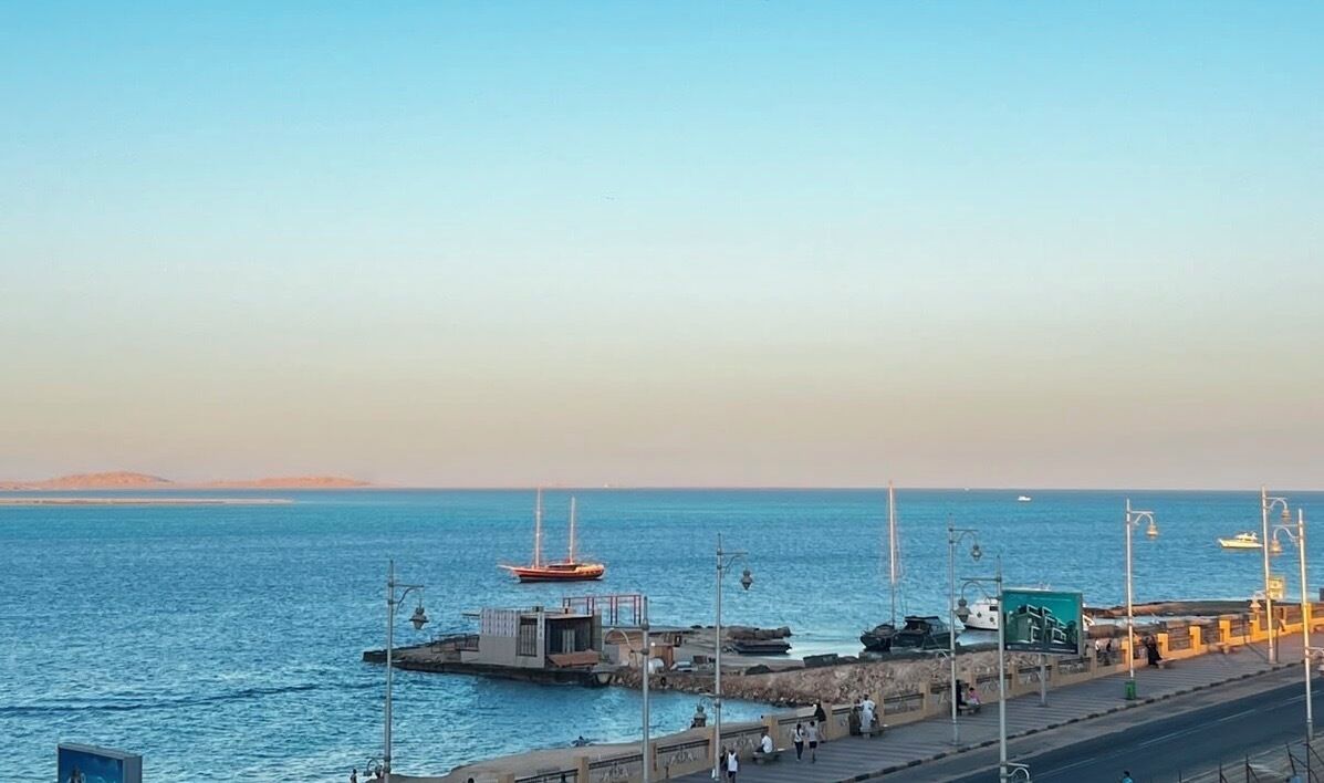 Un muelle costero con un pequeño barco amarrado, una carretera que bordea el agua y un cielo despejado.