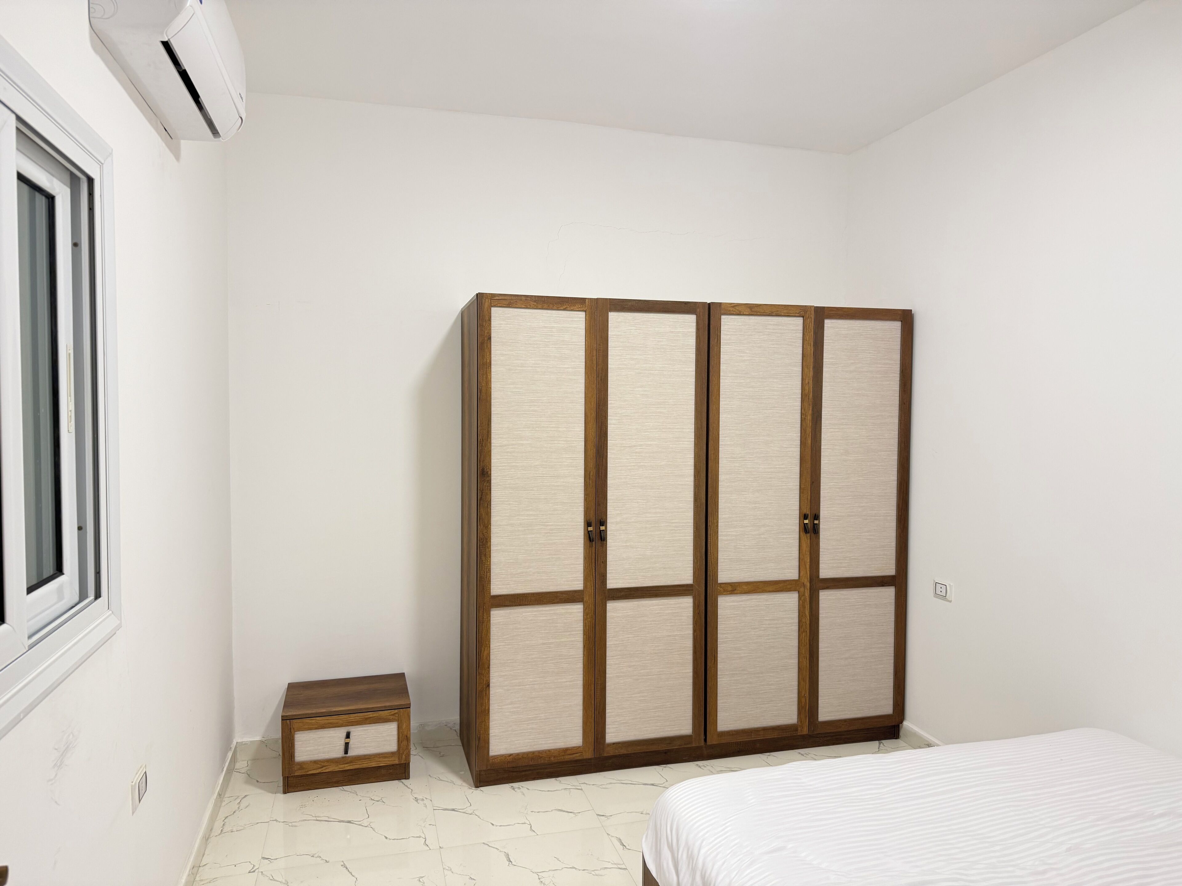 Ein Schlafzimmer mit einem Bett, einem Kleiderschrank, einer kleinen Holzkiste und einem Fenster.