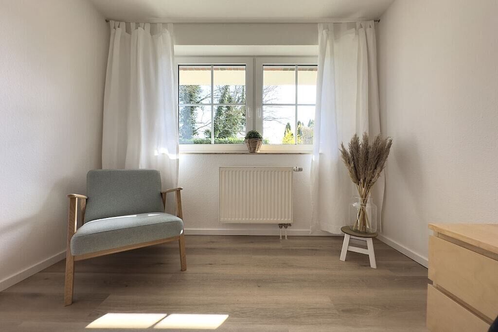 Een kamer met een raam, een radiator, een stoel, een klein tafeltje met een plant en een houten dressoir.