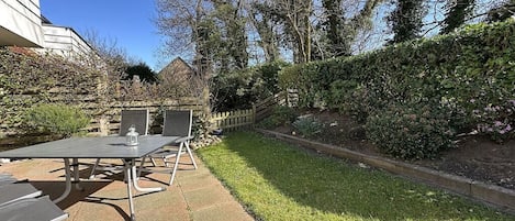 Un espacio al aire libre con patio, una mesa y sillas, rodeado por un seto y un muro de piedra.