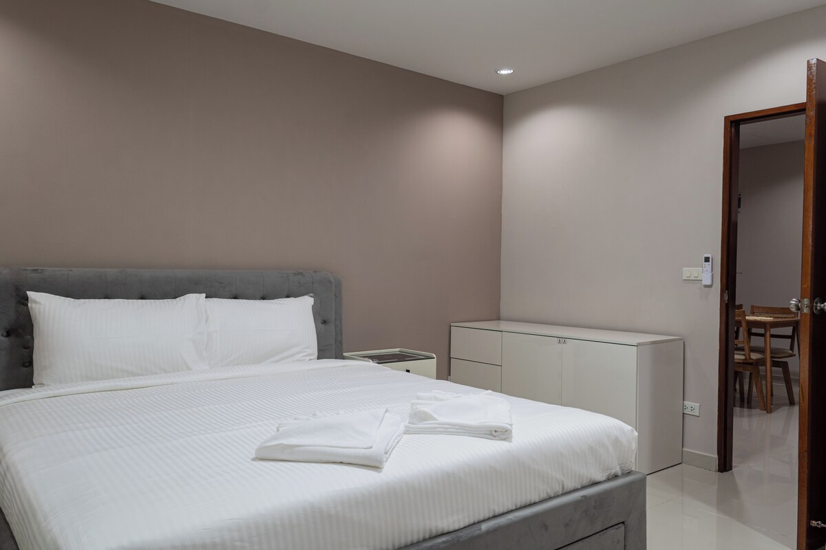 A bedroom with a bed, a bedside table, a wardrobe, and a door leading to a dining area.
