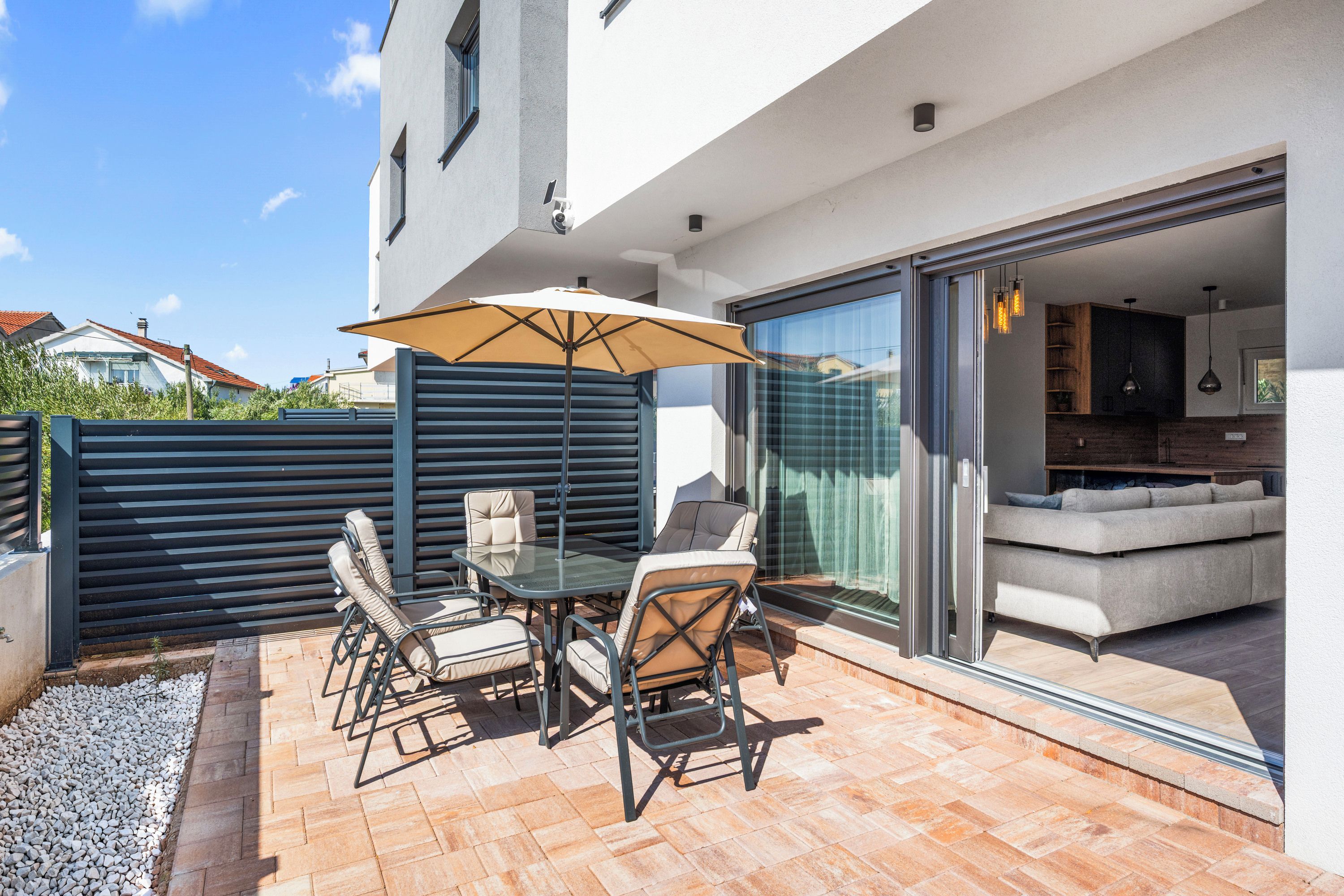 Une terrasse extérieure avec une table et des chaises, un grand parasol et une porte-fenêtre coulissante donnant sur un espace de vie.