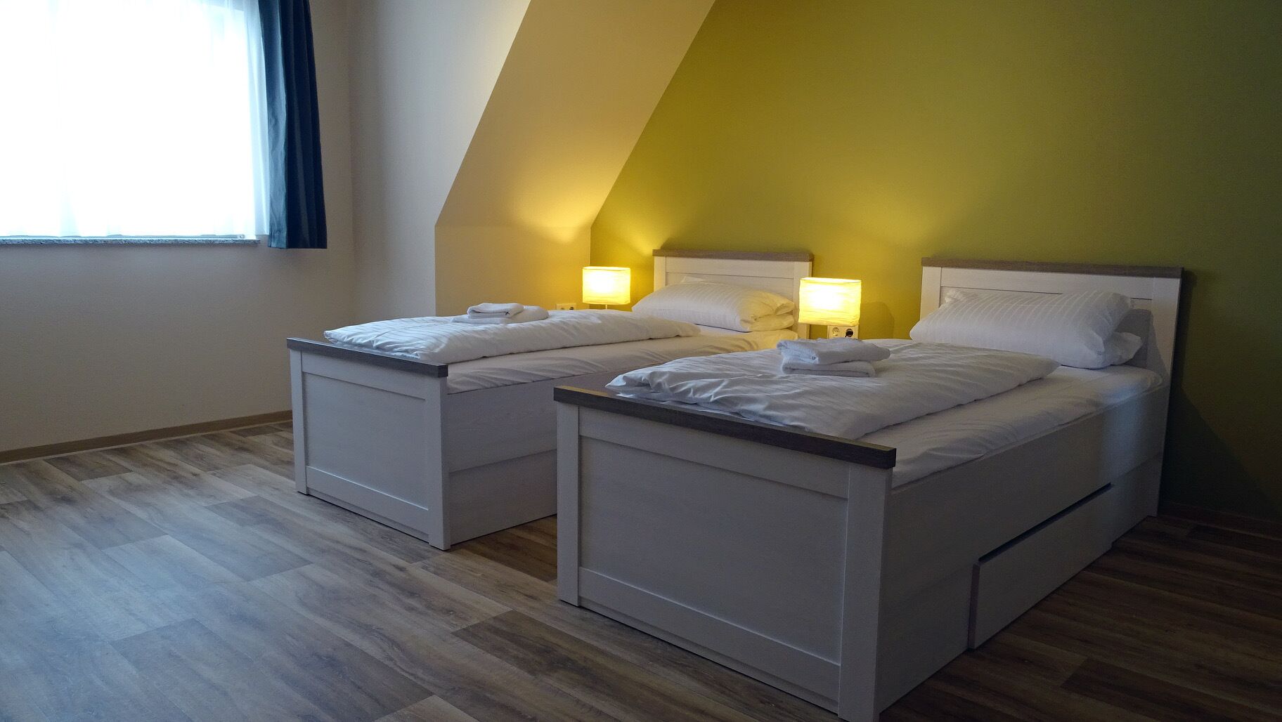 A room with two beds, each with a nightstand and a lamp. The room has wooden flooring and a sloped ceiling.