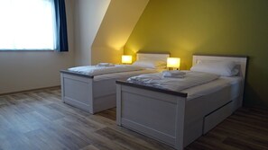 Quarto com duas camas, cada uma com uma mesa de cabeceira e uma luminária. O quarto possui piso de madeira e teto inclinado.