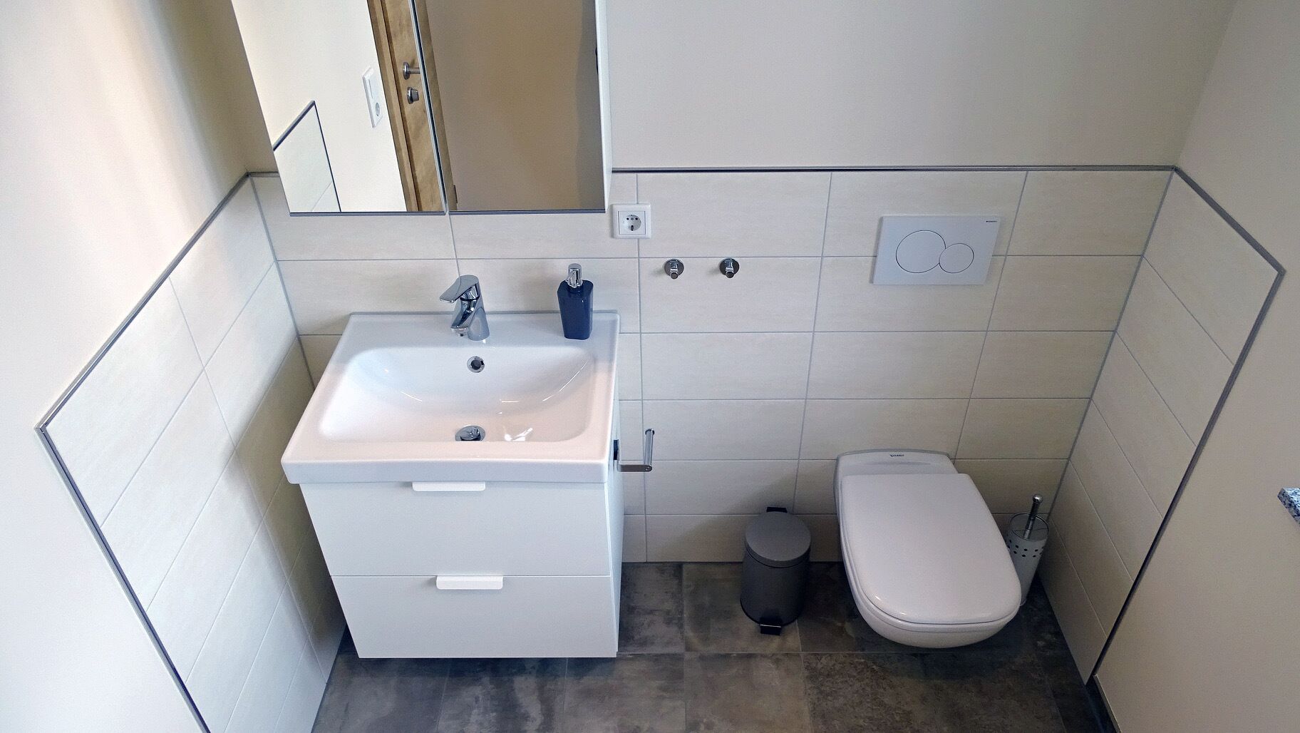 A compact bathroom with a sink, mirror, toilet, and trash bin.