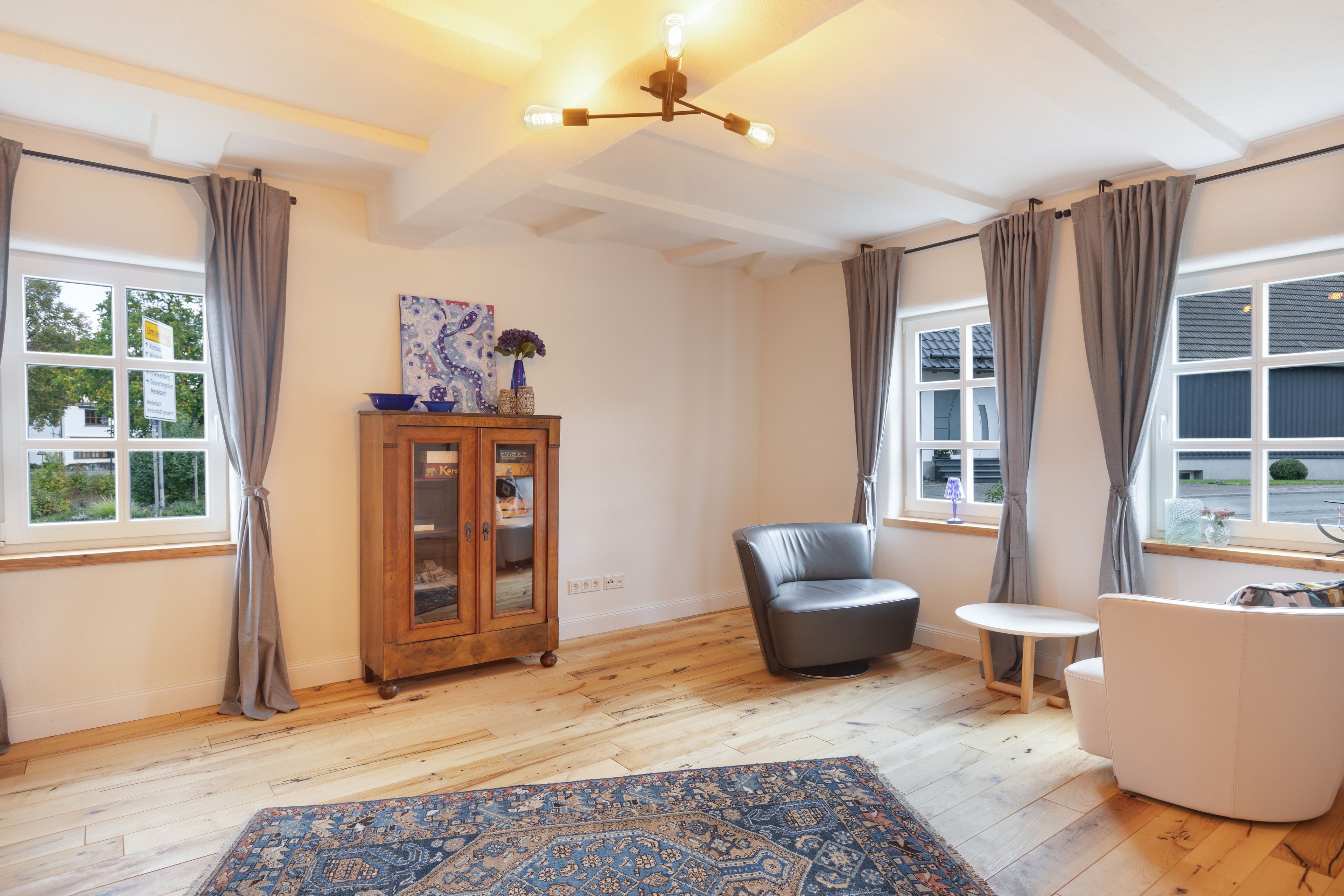 Ein Zimmer mit Holzboden, einem blauen Teppich mit Muster, einem Schrank mit Glastüren, einem grauen Ledersessel, einem weißen Sessel und einem kleinen runden Tisch.