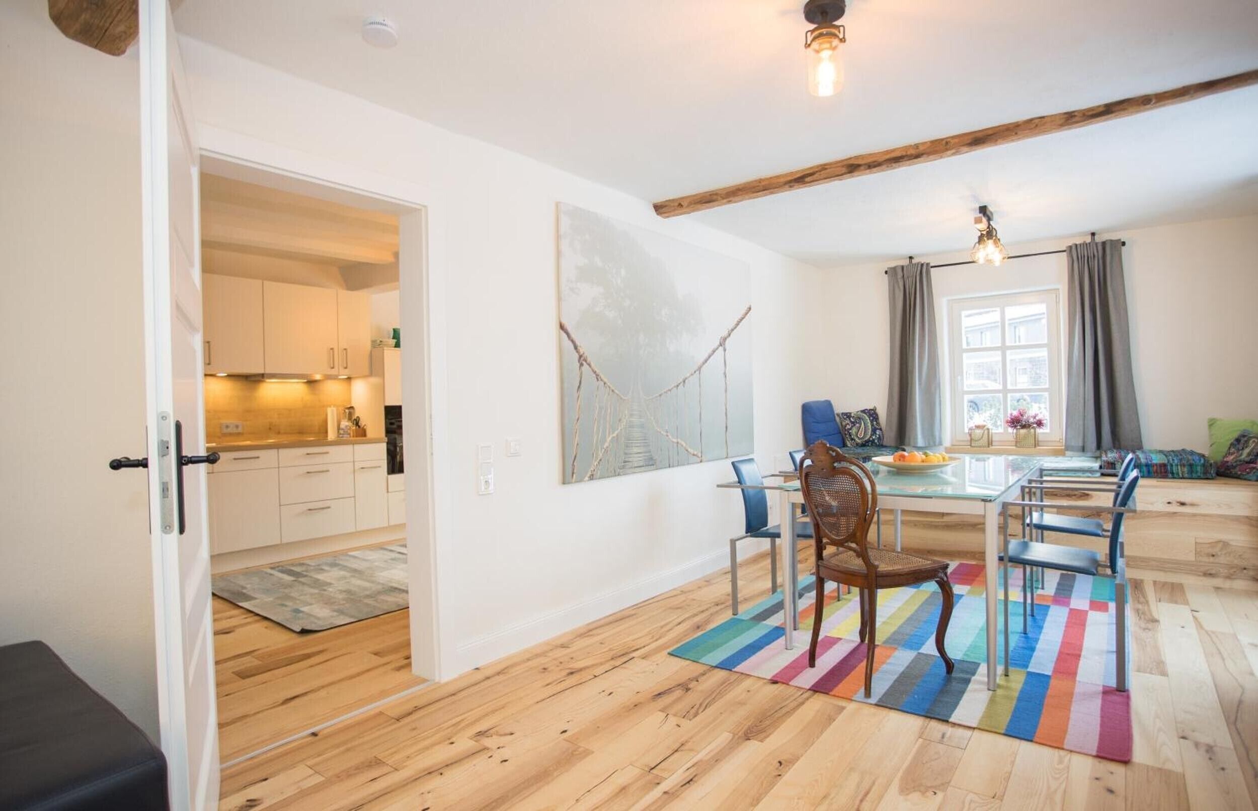 Een moderne keuken met witte kasten, een eethoek met een glazen tafel en stoelen, en een kleurrijk vloerkleed.