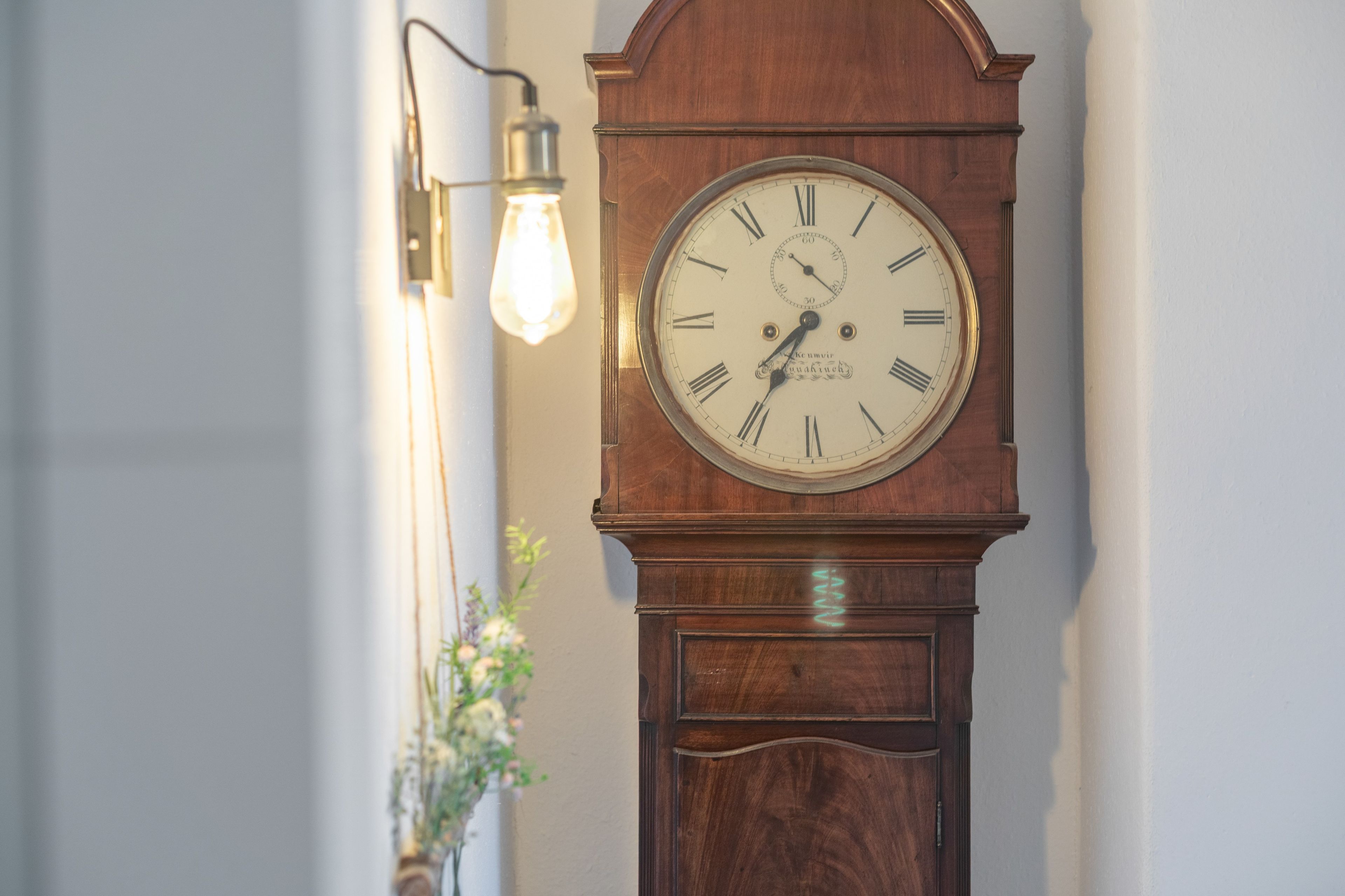Un reloj de pie antiguo fijado a la pared, con números romanos y un péndulo, ubicado junto a una lámpara también fijada a la pared.