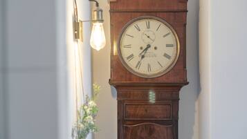 Un reloj de pie antiguo fijado a la pared, con números romanos y un péndulo, ubicado junto a una lámpara también fijada a la pared.