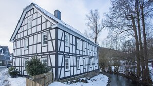 A half-timbered house with a river in front, wooden fence, and snow on the ground. - Apartment 'Denkmal Haus Hugo EG' with Private Terrace, Shared Garden and Wi-Fi (Medebach)