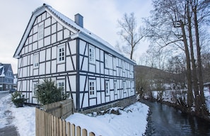 Ein Fachwerkhäuschen mit einem Fluss davor, einem Holzzaun und Schnee auf dem Boden.