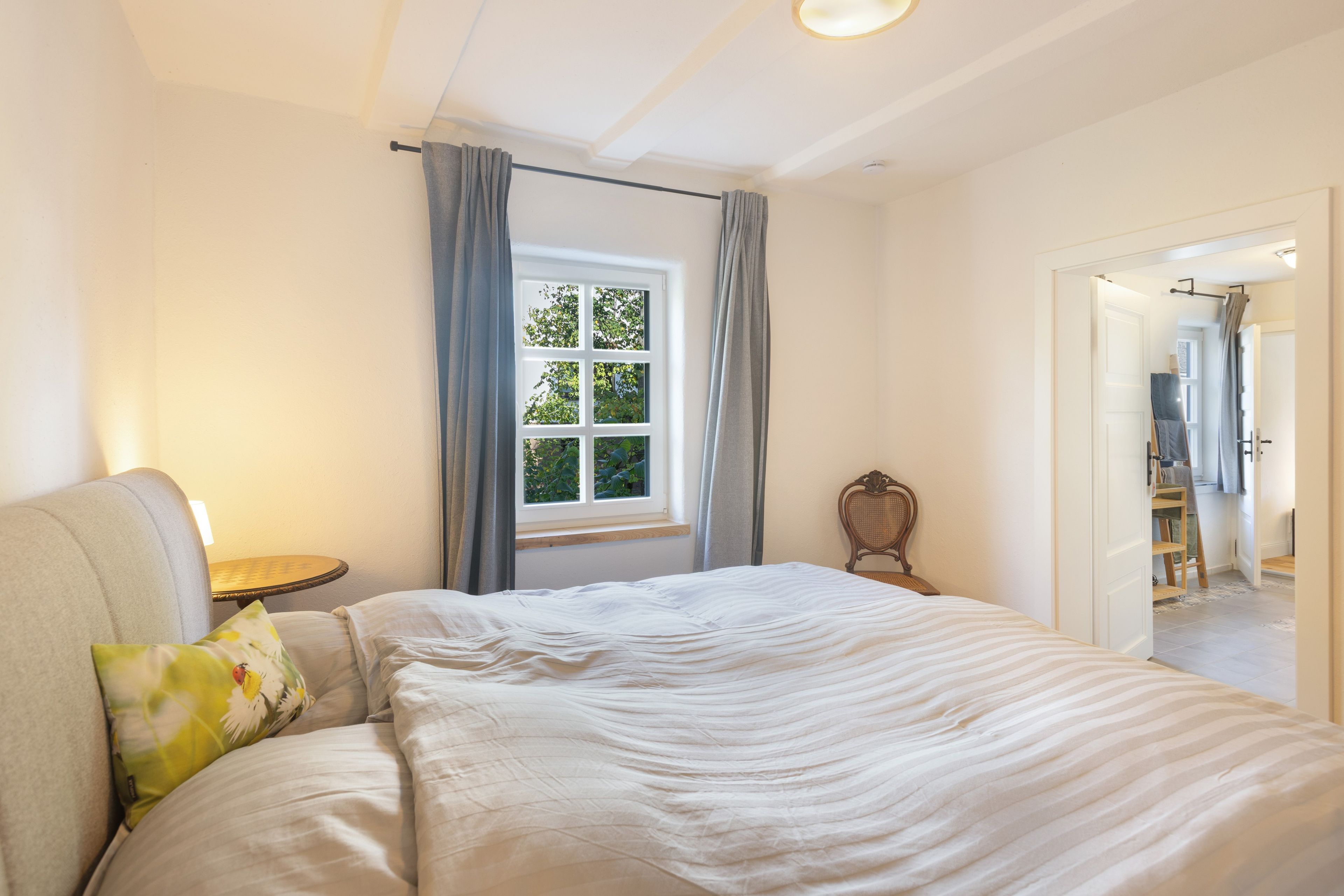 A bedroom with a bed, a nightstand, a chair, a window with curtains, and a door leading to another room.