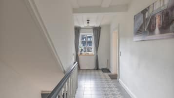A hallway with a tiled floor, a window with curtains, a painting on the wall, and a door at the end.