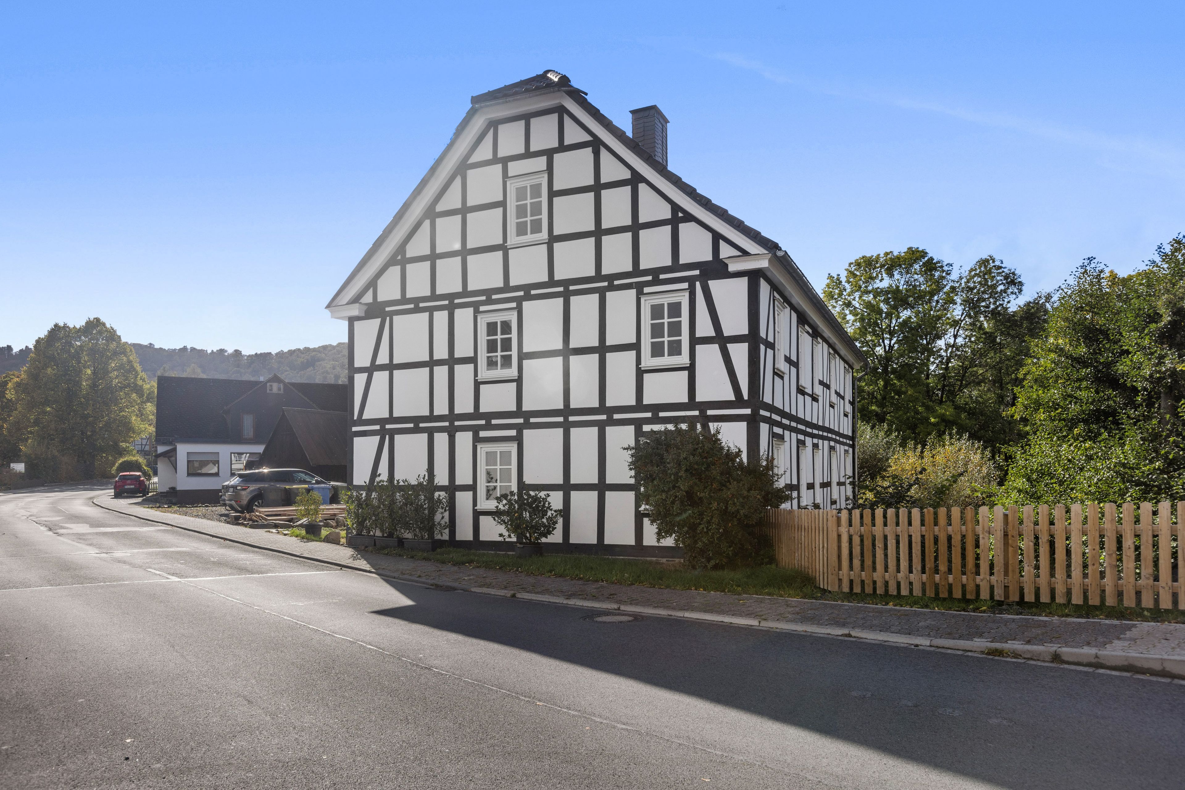 Ein traditionelles Fachwerkhäuschen mit geneigtem Dach, einem umzäunten Garten und einer Straße im Vordergrund.