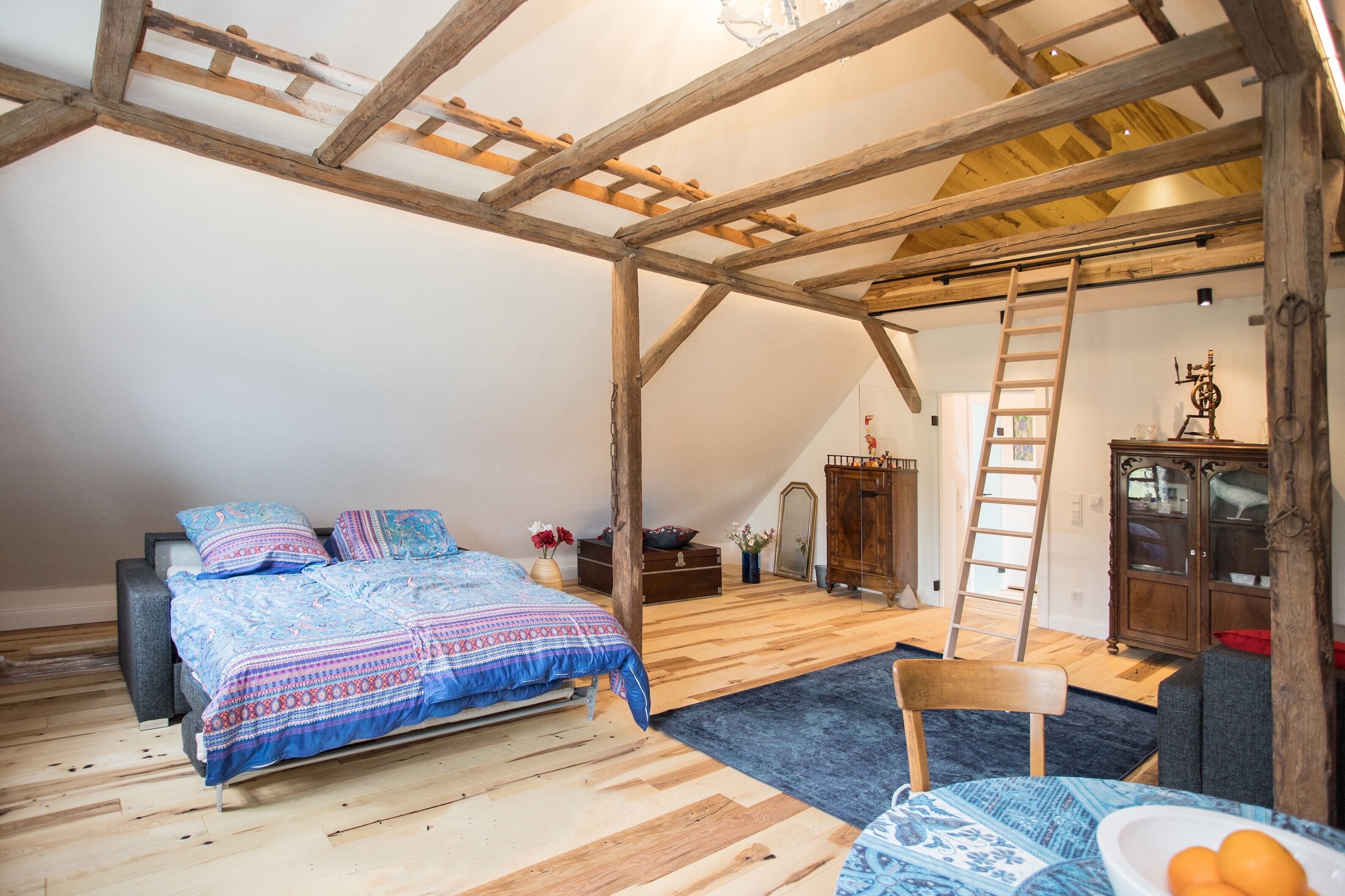Quarto com teto inclinado, cama com roupa de cama estampada, cômoda de madeira, cadeira e escada.