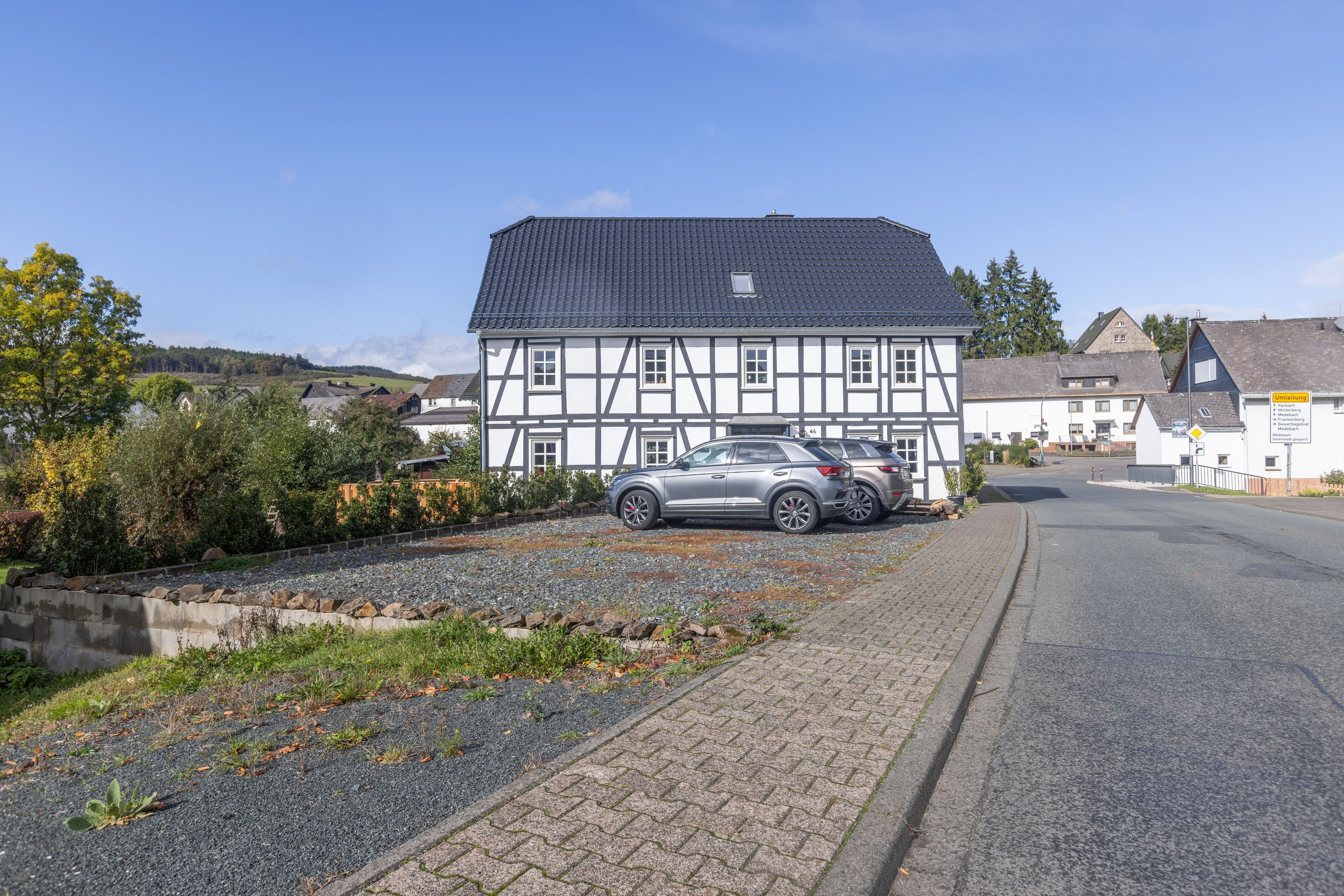 Ein traditionelles Fachwerkhäuschen mit einem geparkten Auto davor, einer Schotterauffahrt und blauem Himmel.