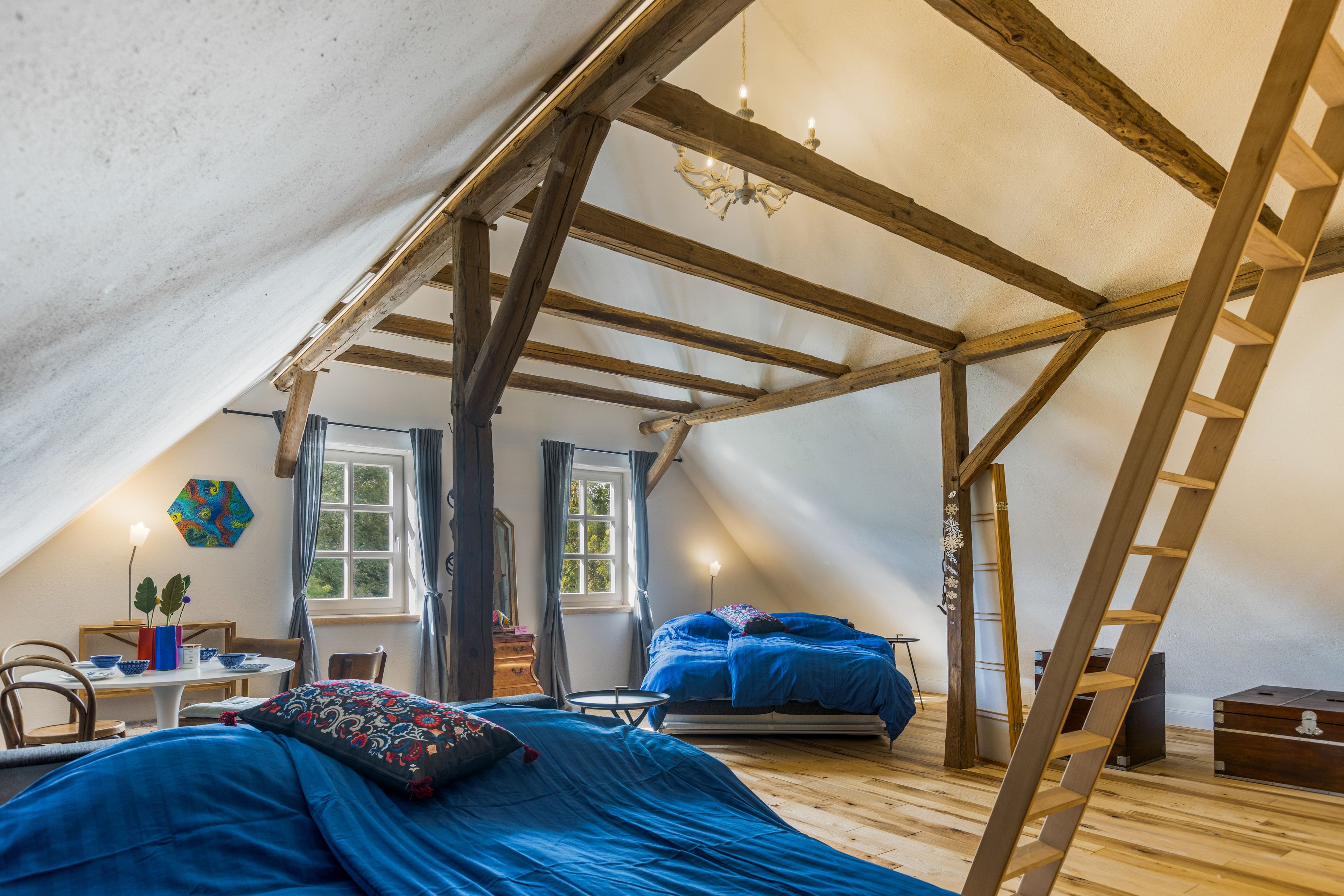 A loft bedroom with a sloped ceiling, wooden beams, a bed with blue bedding, a dresser, a chair, and a window.