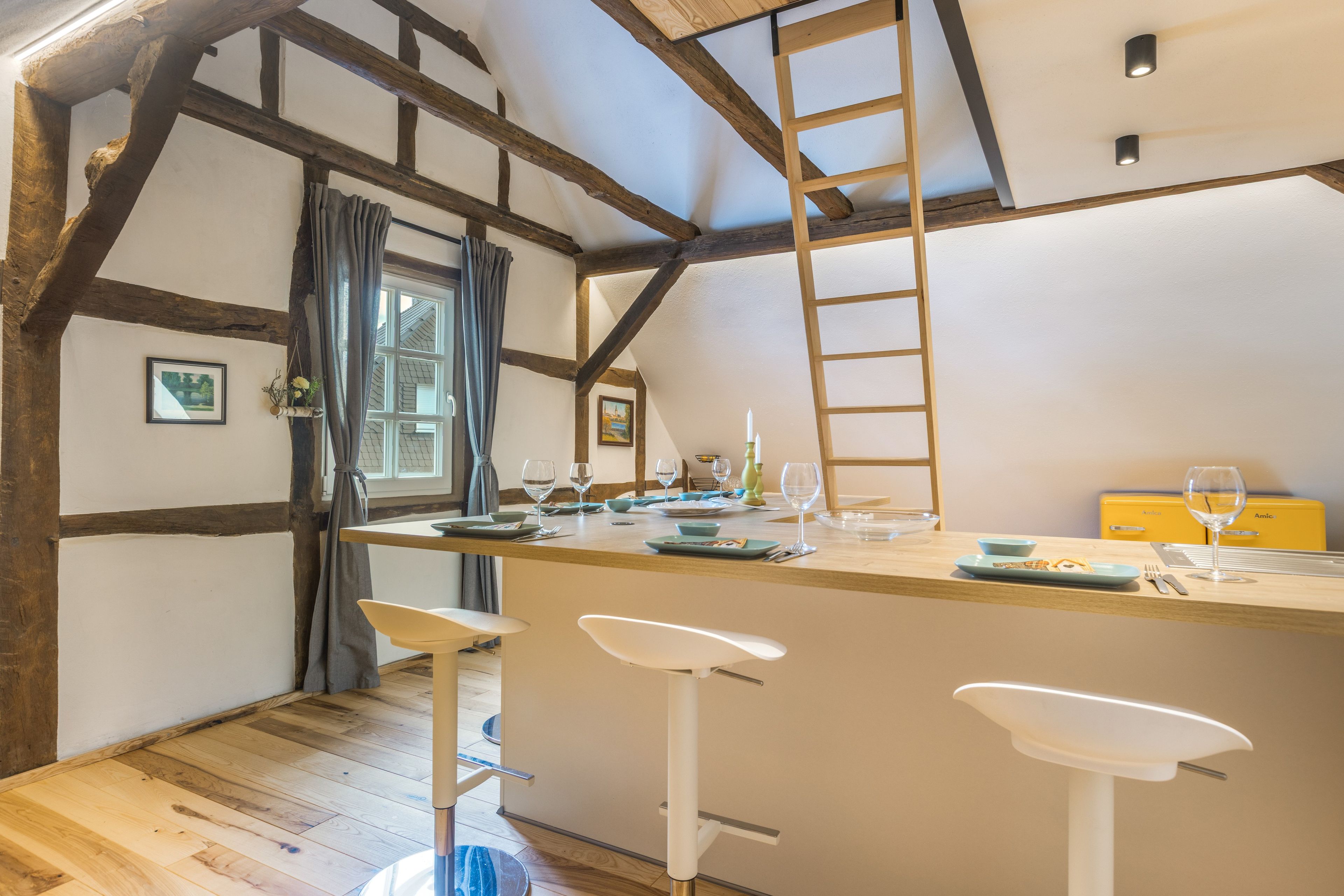 A rustic kitchen with exposed wooden beams, a central island with bar stools, and a window with curtains.