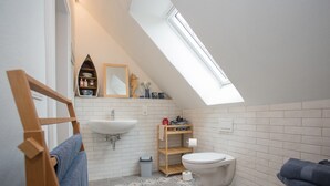 Une petite salle de bain moderne avec un puits de lumière, des carreaux blancs, une étagère en bois où sont rangés les articles de toilette, un lavabo blanc et des toilettes.