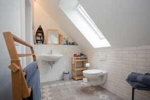 A small, modern bathroom with a skylight, white tiles, a wooden shelf with toiletries, a white sink, and a toilet. - Denkmal Haus Hugo Loft Apartment with Alpine Flair and Wi-Fi (Medebach)