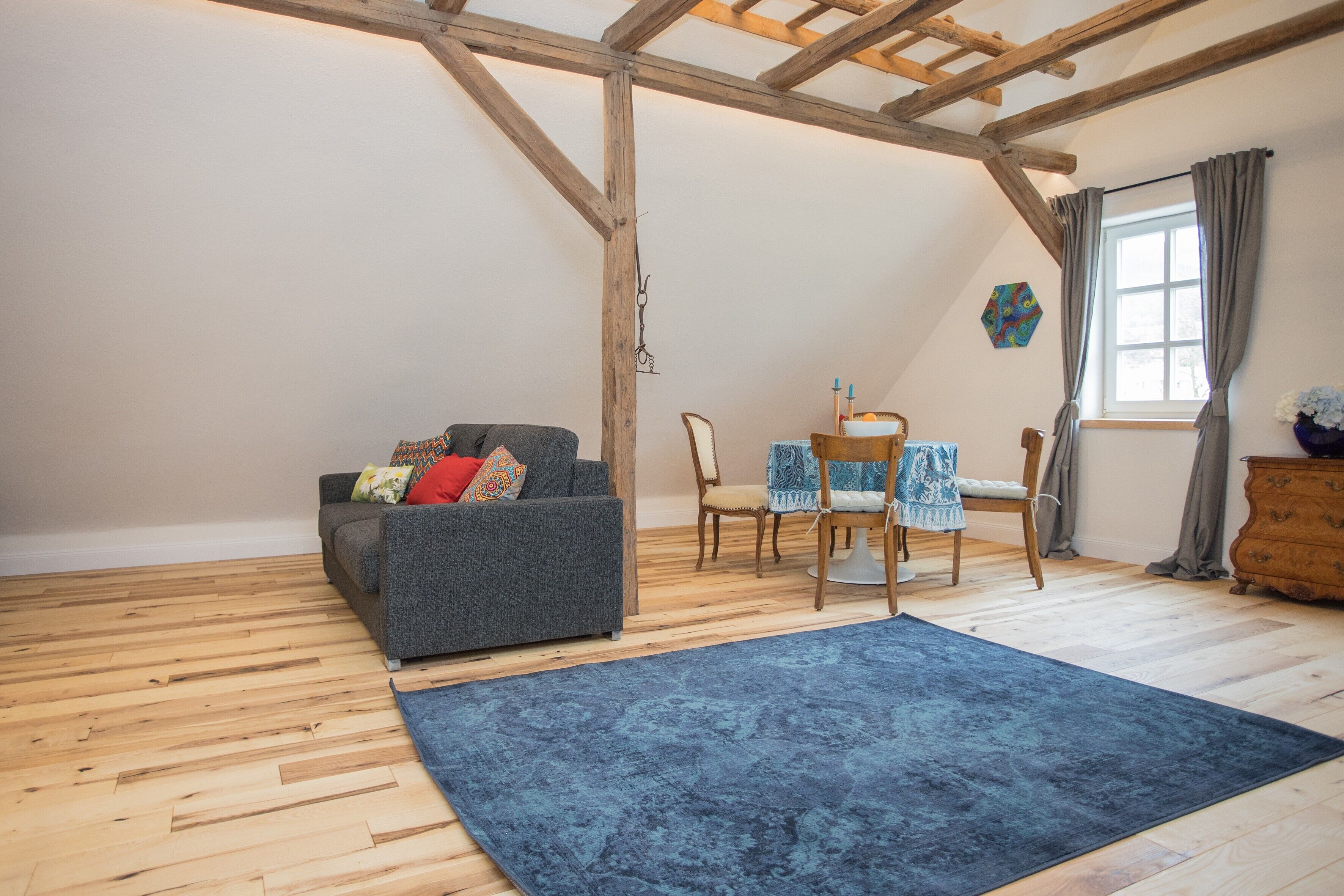 Um quarto estilo loft com vigas de madeira aparentes, um sofá cinza com almofadas estampadas, uma mesa de jantar com cadeiras e um tapete azul.