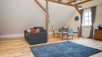 Um quarto estilo loft com vigas de madeira aparentes, um sofá cinza com almofadas estampadas, uma mesa de jantar com cadeiras e um tapete azul.
