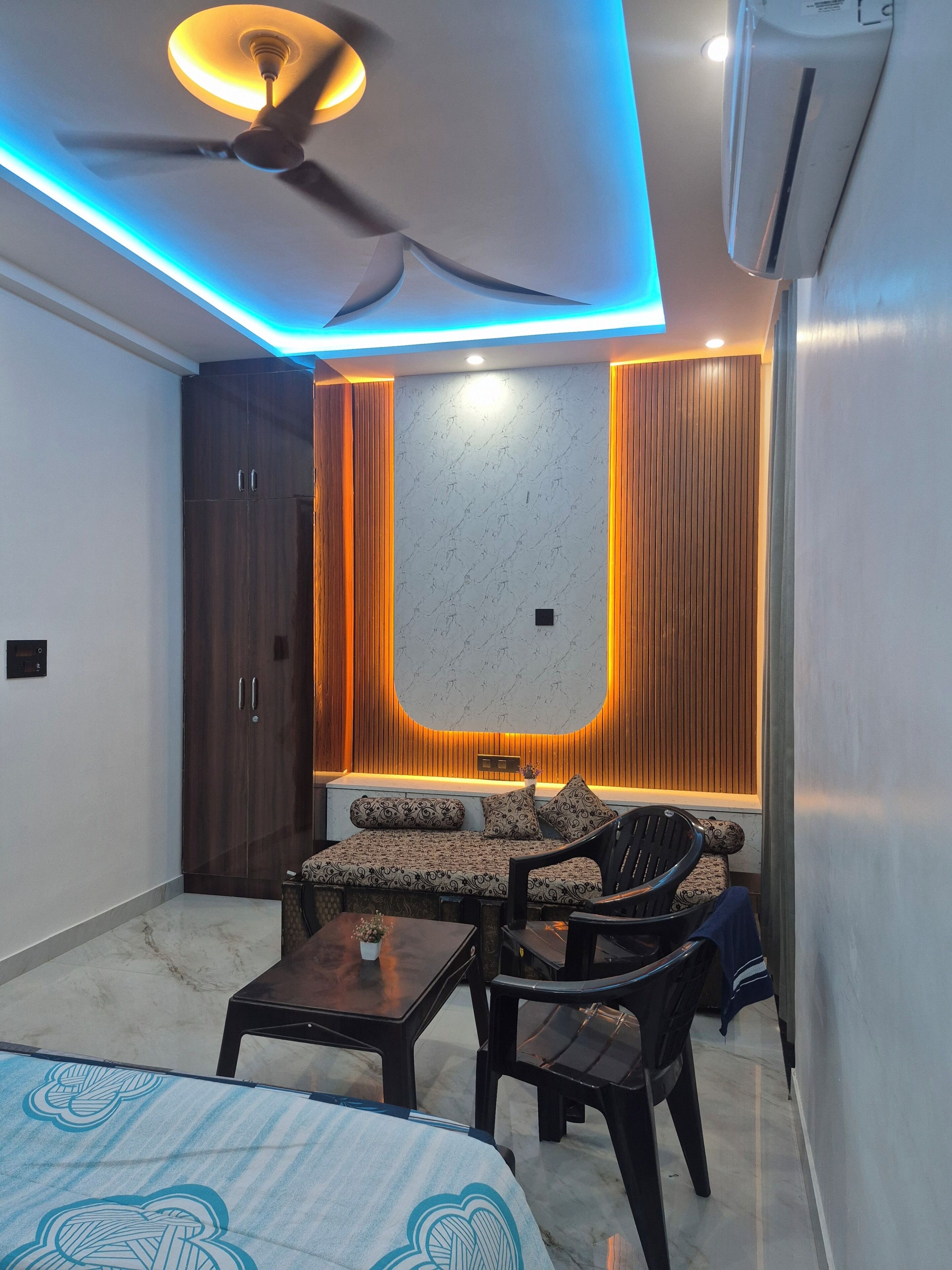 Una habitación compacta con cama, sofá, sillón y una pequeña mesa. Cuenta con ventilador de techo, unidad de aire acondicionado y una lámpara fijada a la pared.