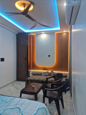 A compact room with a bed, a sofa, a chair, and a small table. There is a ceiling fan, an air conditioning unit, and a wall-mounted light fixture. - 1BHK Flat | Near Jaipur Airport (Jaipur)