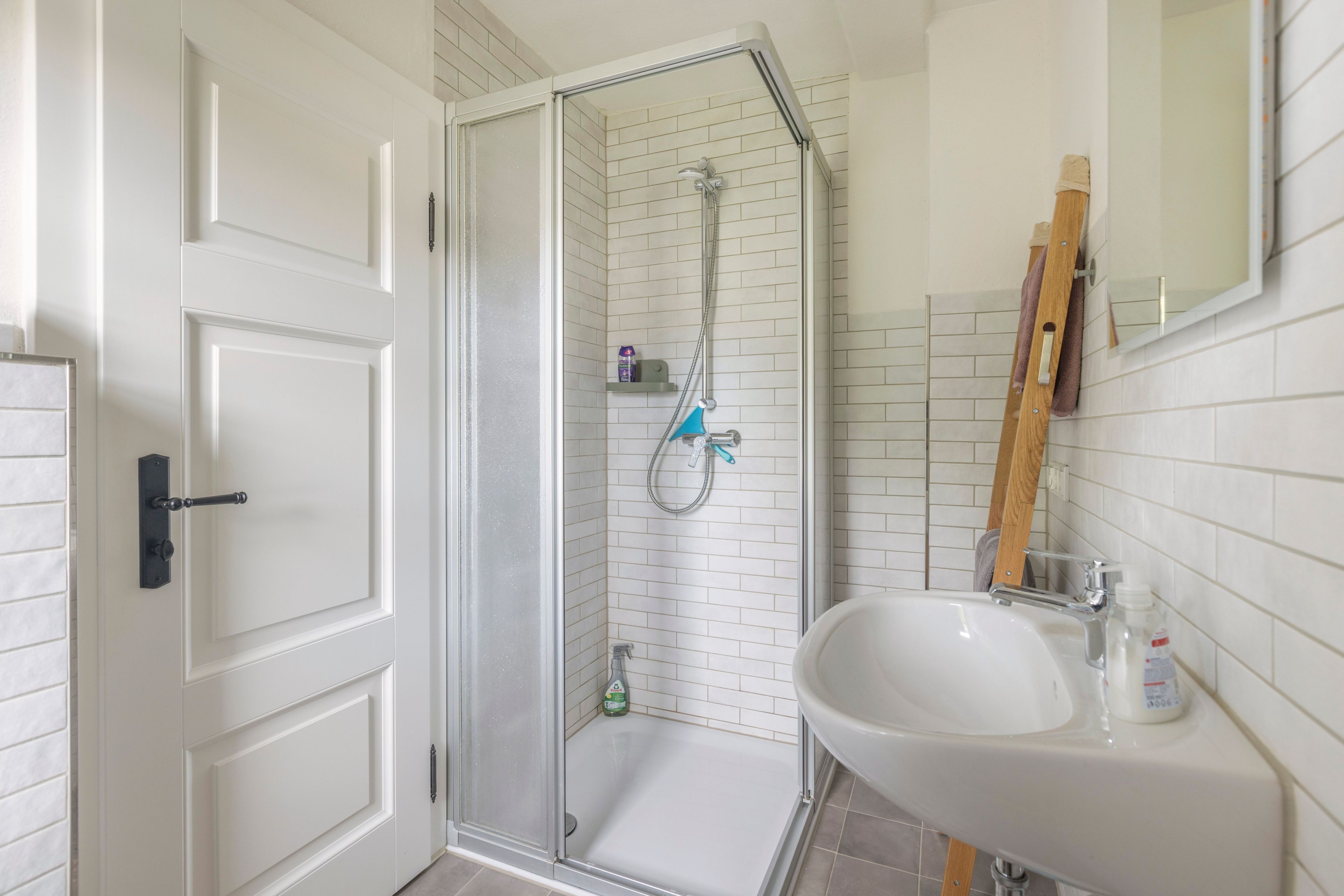 Ein Badezimmer mit weißer Tür, einer Dusche mit Glastür, einem weißen Waschbecken und einem Spiegel.