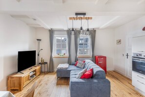 Un salon moderne avec un canapé gris, un téléviseur à écran plat, une table d’appoint en bois et un réfrigérateur rouge de la marque Amica.
