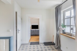 Un couloir mène à un salon équipé d’un canapé, d’un téléviseur et d’une fenêtre avec des rideaux.
