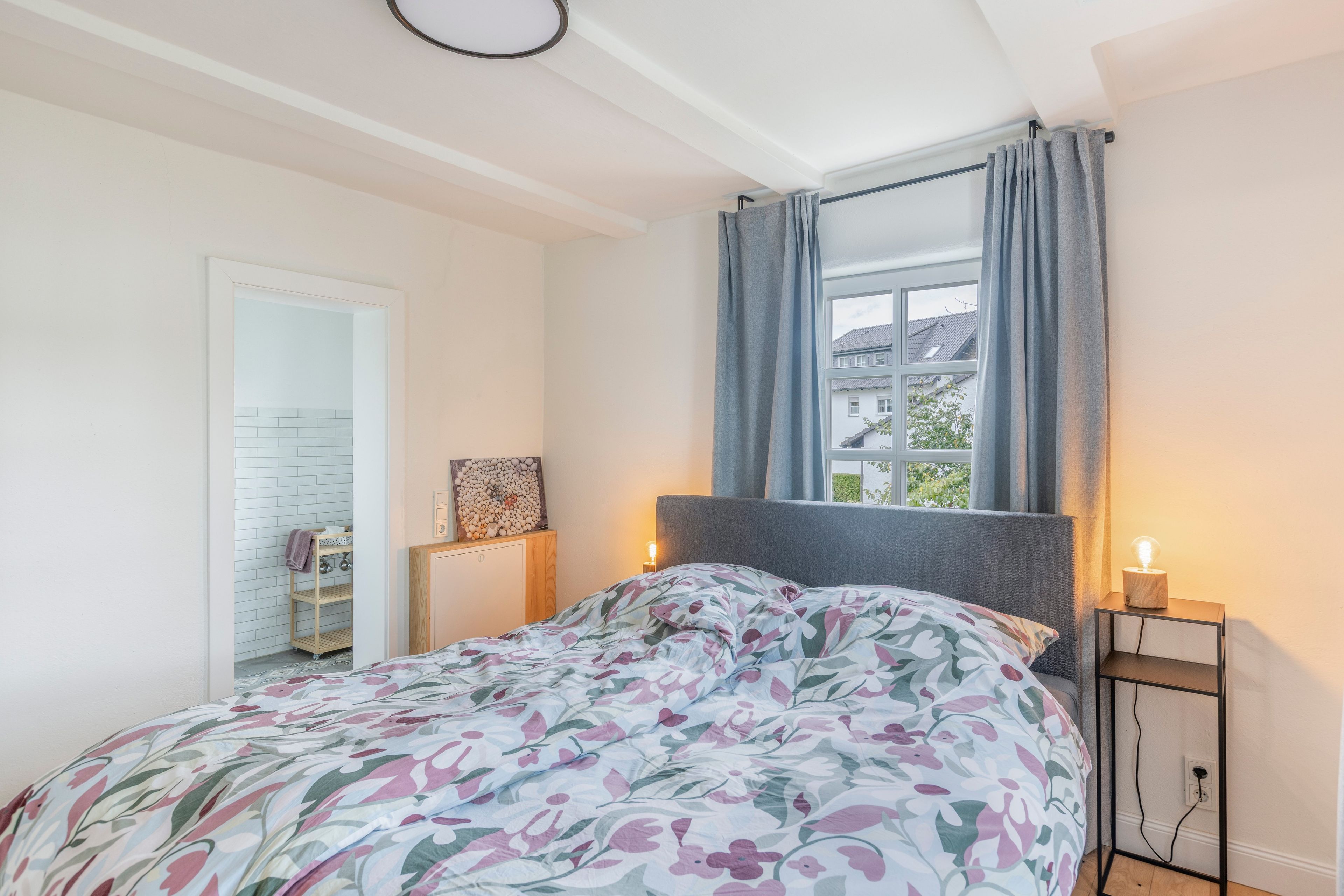 Een slaapkamer met een bed, nachtkastje, raam met gordijnen en uitzicht op gebouwen.