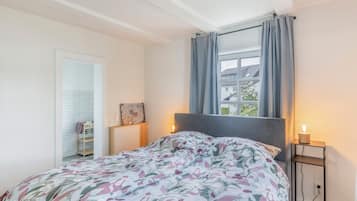 Een slaapkamer met een bed, nachtkastje, raam met gordijnen en uitzicht op gebouwen.