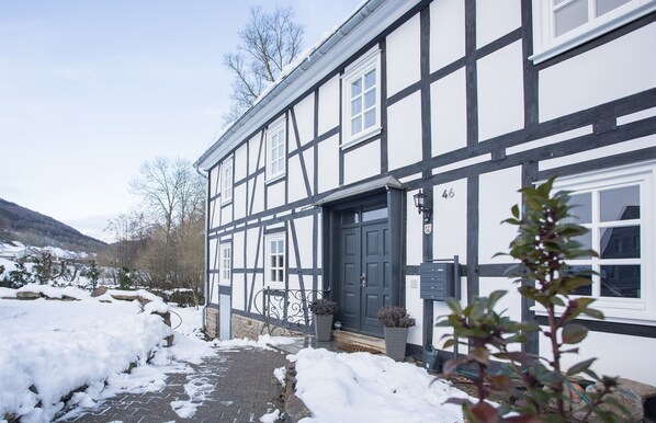 A traditional half-timbered house with a dark entrance door, white windows, and a snow-covered pathway leading to the entrance.