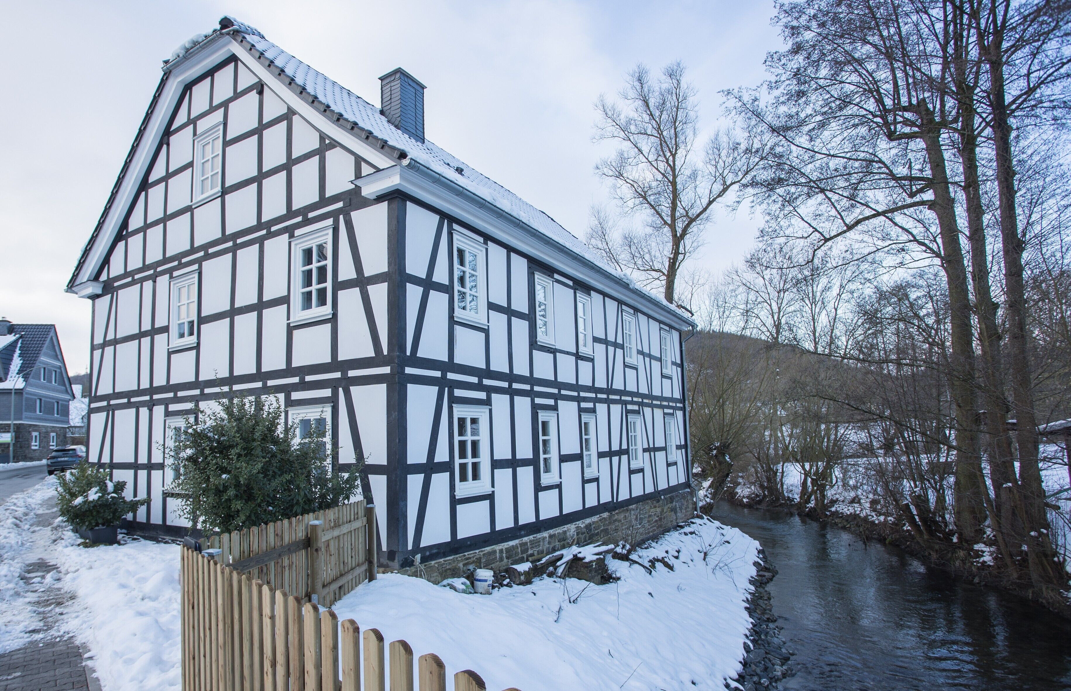 Een vakwerkhuis met een rivier ervoor, houten omheining en sneeuw op de grond.