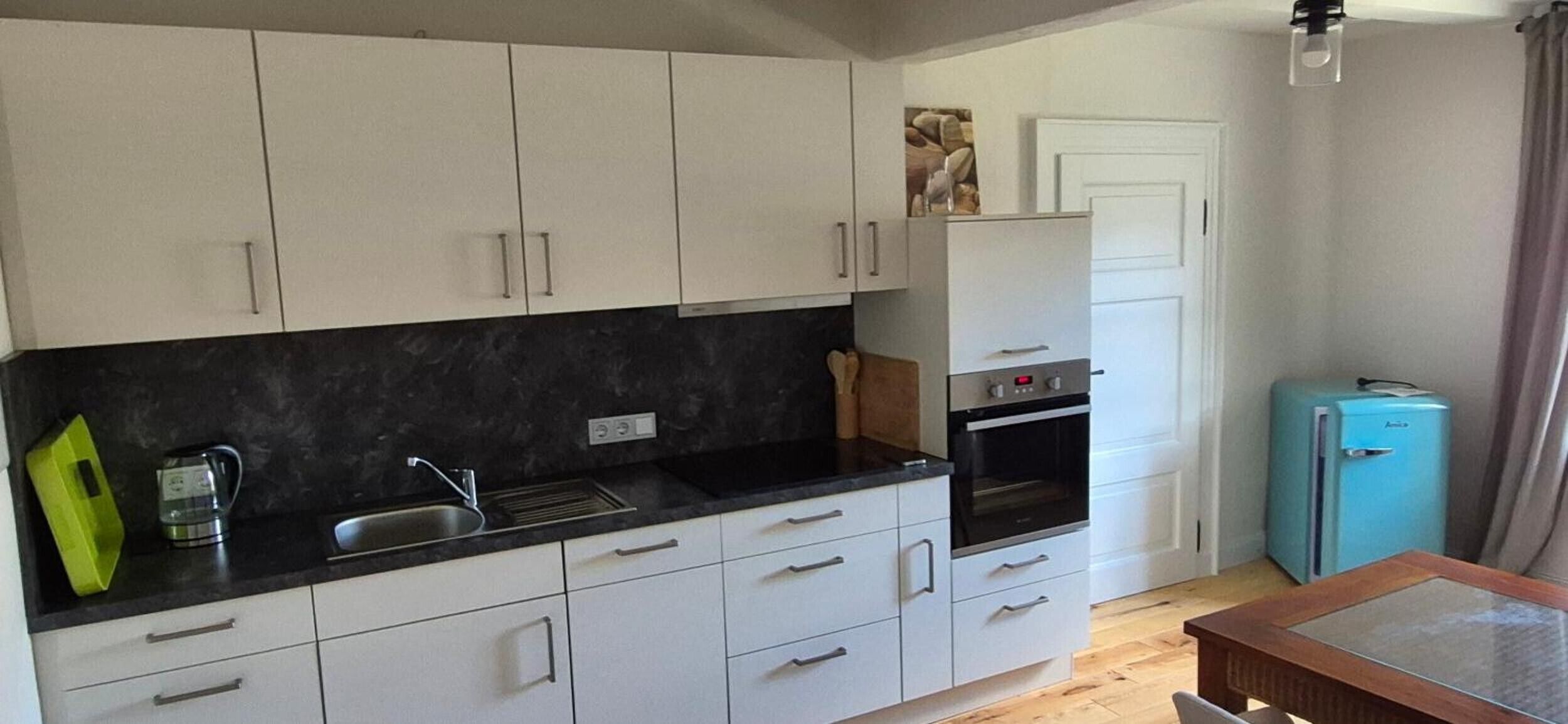 Eine kompakte Küche mit weißen Schränken, einer schwarzen Arbeitsplatte, einem Spülbecken und einem kleinen, blauen Kühlschrank.