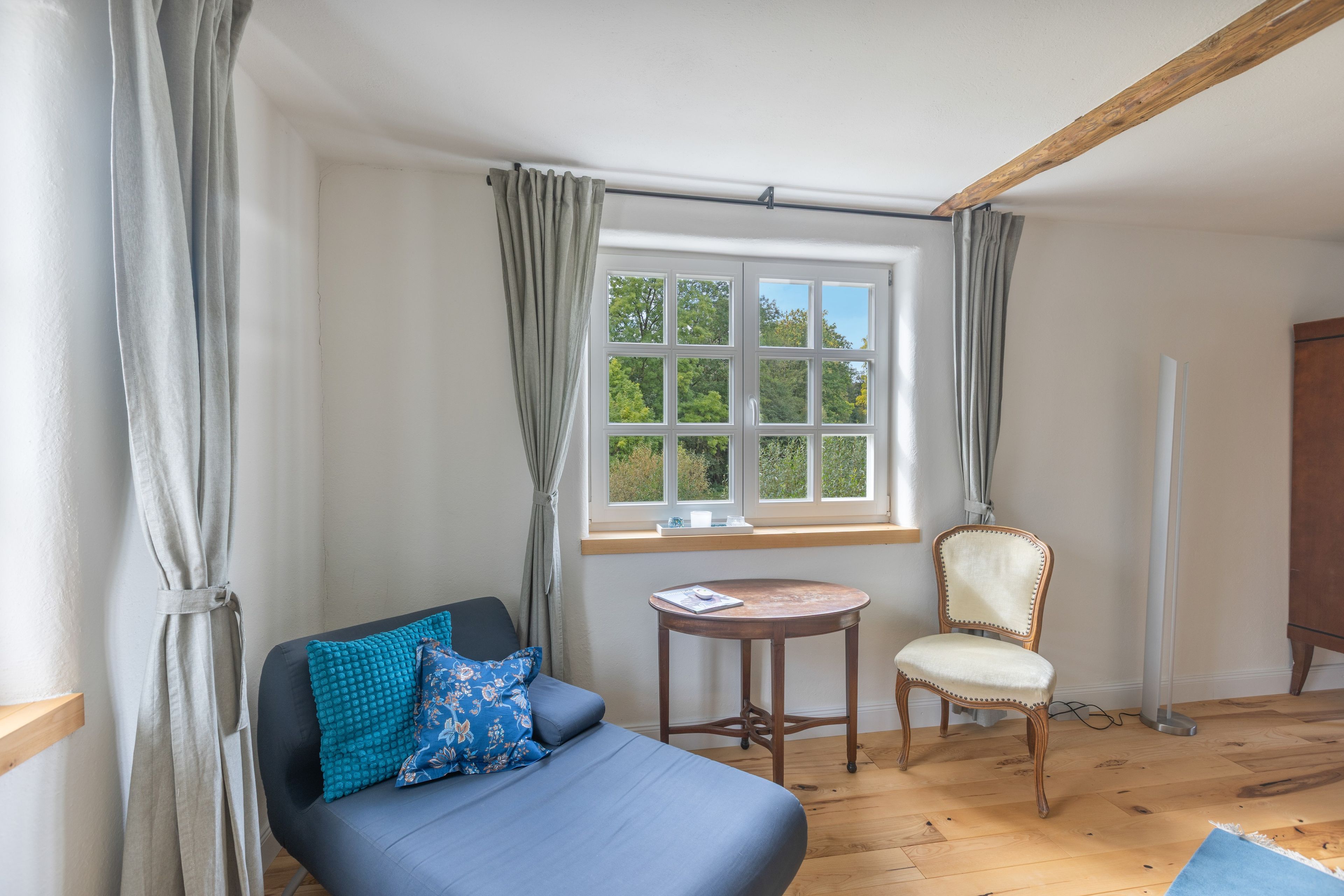 Ein Schlafzimmer mit einem Bett, einem Sessel, einem kleinen Tisch und einem Fenster mit Vorhängen.