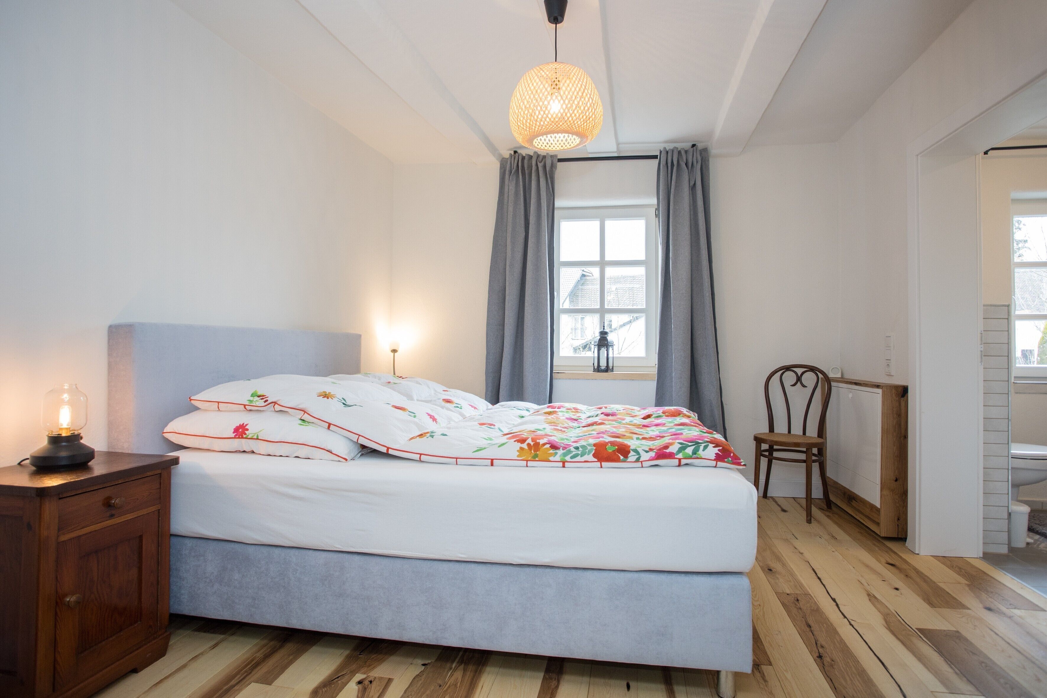 Een slaapkamer met een bed, nachtkastje, stoel, raam met gordijnen en een hanglamp.