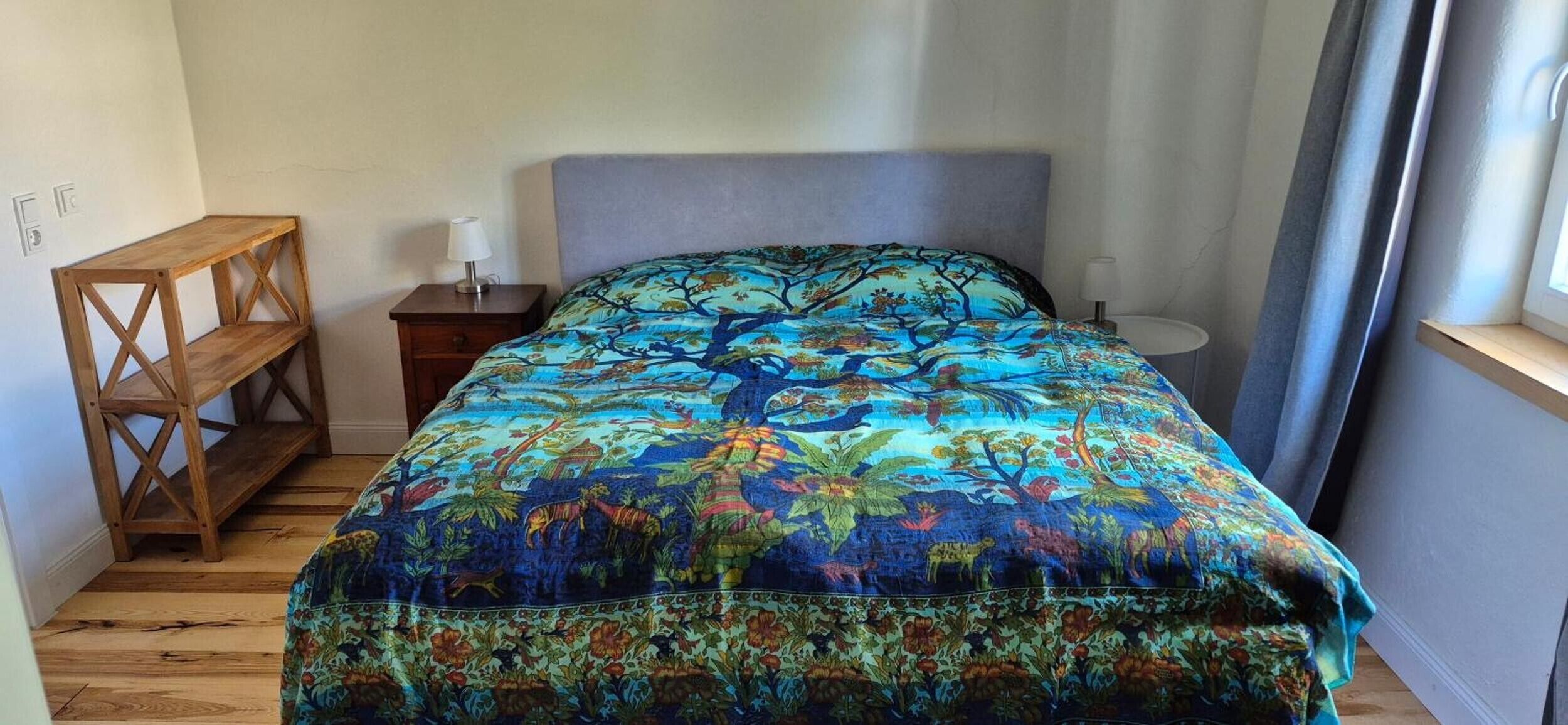 Quarto com cama, mesa de cabeceira de madeira, abajur e janela com cortinas.