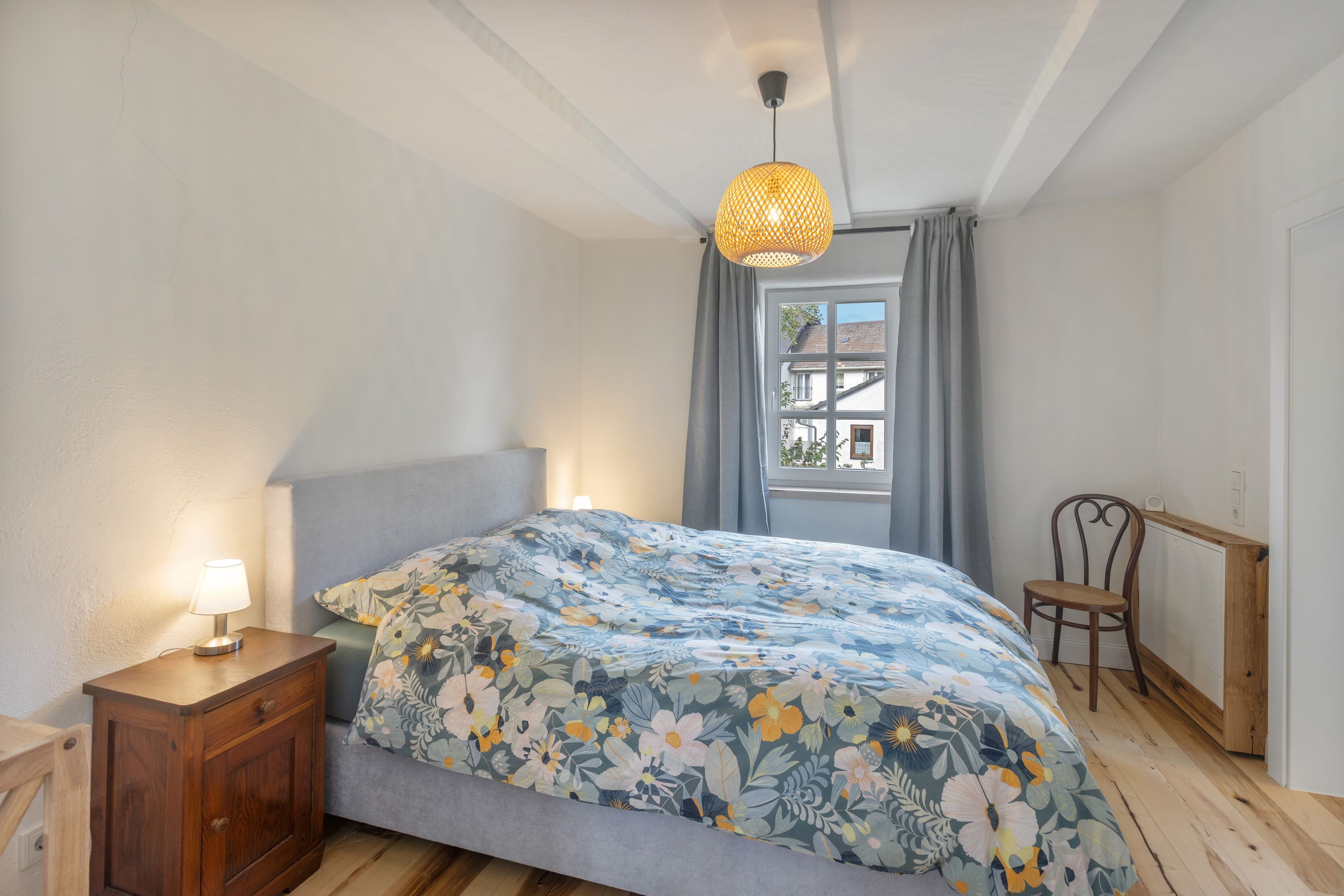 Ein Schlafzimmer mit Bett, Nachttisch, Sessel, Fenster und einer Pendelleuchte.