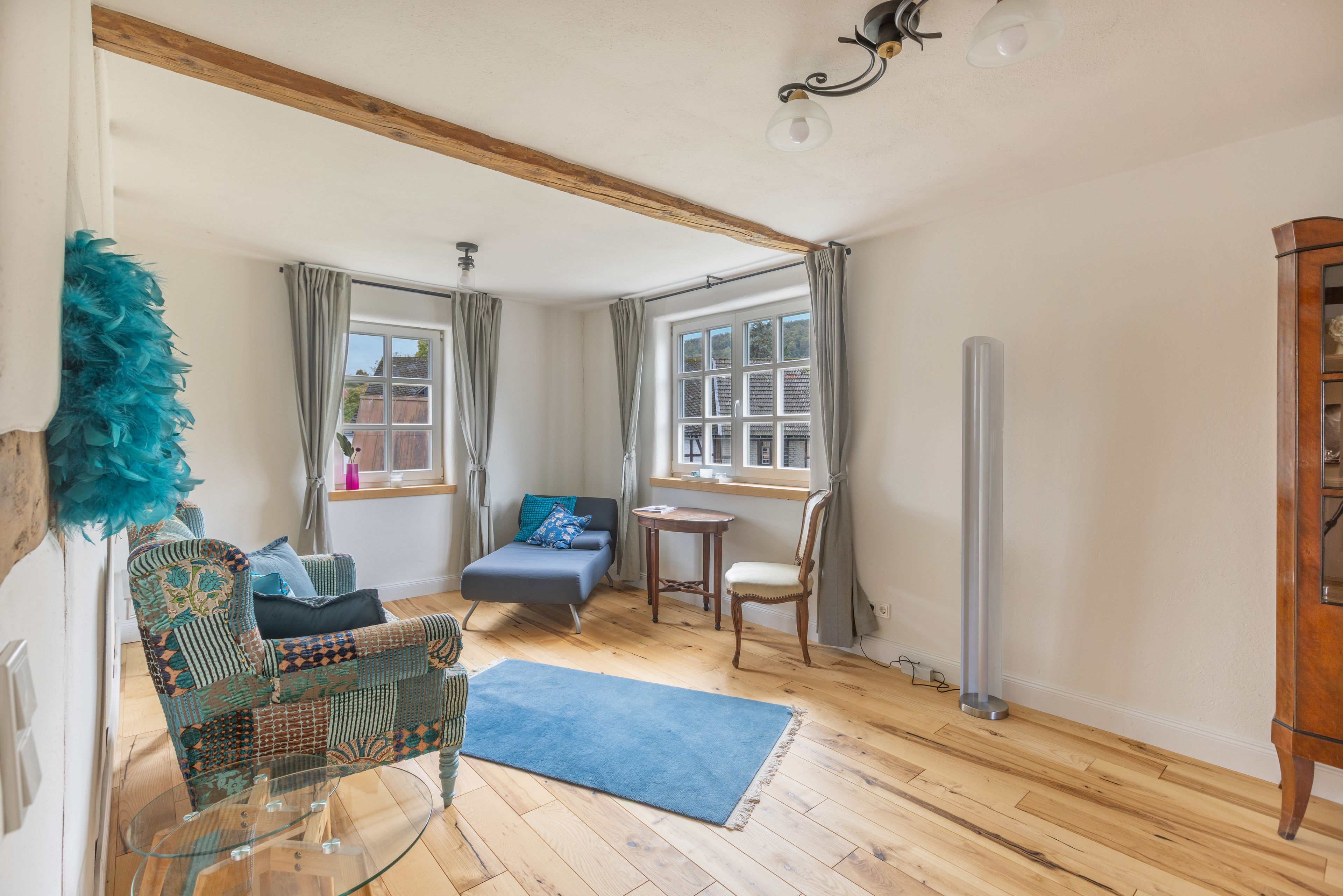 A room with a glass table, a blue rug, a wooden floor, a chair, a sofa, and a window with curtains.