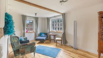 A room with a glass table, a blue rug, a wooden floor, a chair, a sofa, and a window with curtains.