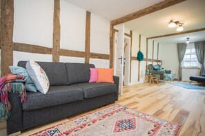 Een gezellige woonkamer met een grijze bank, kleurrijke kussens, een vloerkleed met patroon, houten balken en uitzicht op een andere kamer.