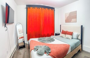 Un dormitori modern amb un llit, una taula de nit, un mirall i un televisor penjat a la paret.