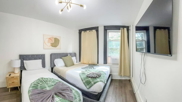 A hotel room with two beds, a bedside table with a lamp, a ceiling light fixture, and a large window with curtains. - 20 Mins to Manhattan w/Full kitchen (Apt. 5) (West New York)