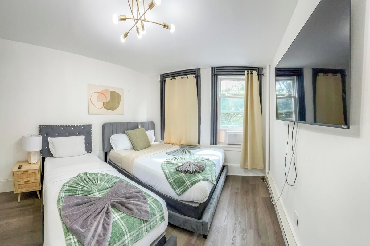 Habitació d'hotel amb dos llits, una taula de nit amb una làmpada, un llum al sostre i una finestra gran amb cortines.