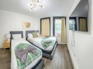 Habitación de hotel con dos camas, una mesita de noche con lámpara, un candelabro en el techo y un amplio ventanal con cortinas.